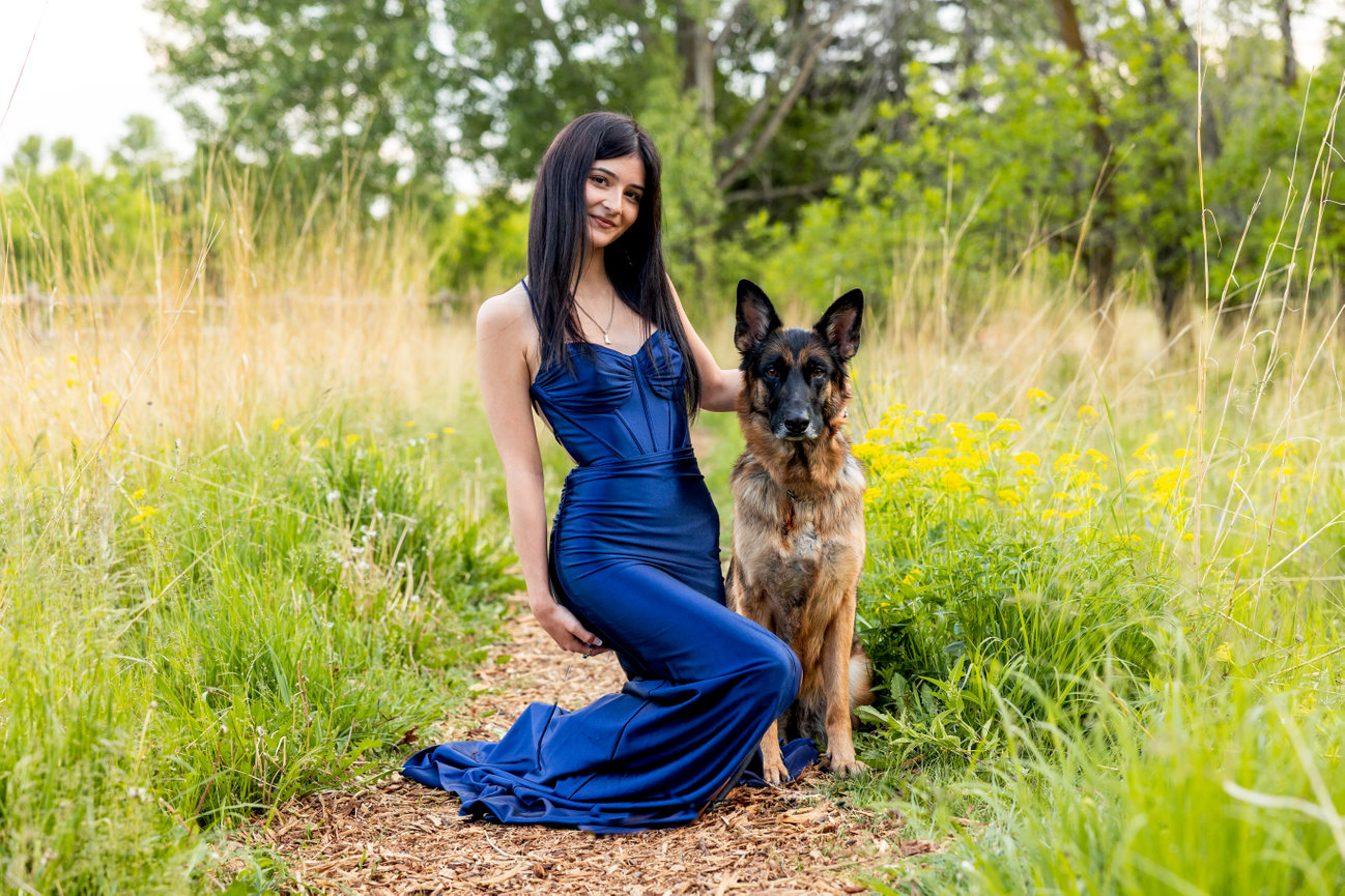 A woman in a blue dress kneels beside a German Shepherd on a grassy path.