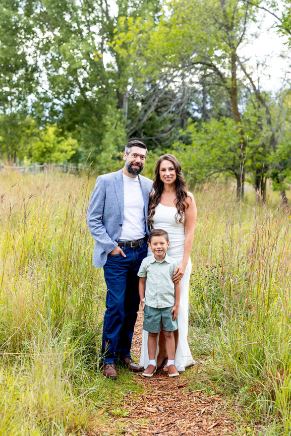 Family posing in a grassy field, with tall trees in the background. Adult wearing jacket, one child, and another adult in a dress.