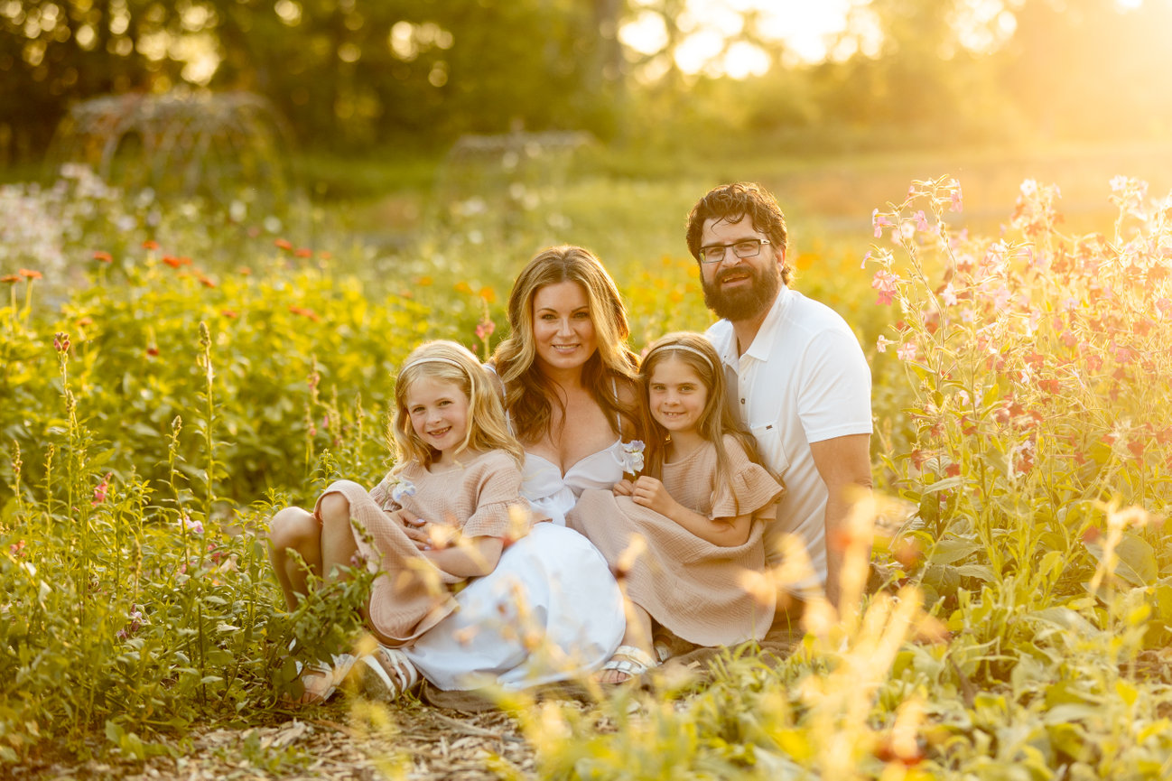 A family of four sits together in a sunlit flower field, surrounded by colorful blooms and lush greenery.