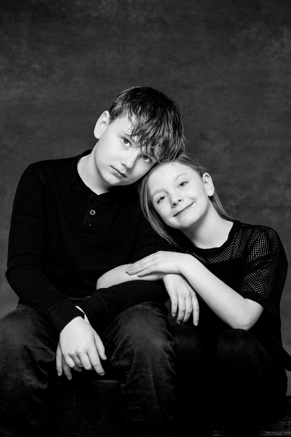 Two children in black clothing sit closely, with one resting their head on the other's shoulder, against a dark background.