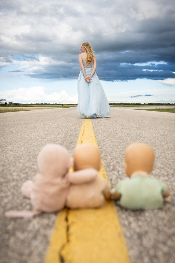 Graduate in a blue dress stands on an airport runway under dramatic clouds, with toy figures in the foreground