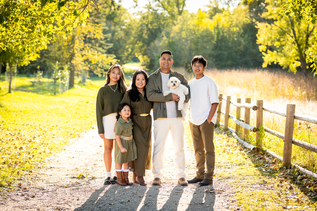 Winnipeg family standing on a sunlit autumn path beside a wooden fence, holding a small dog