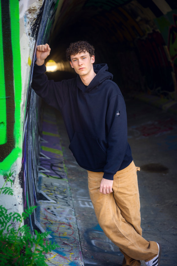 Young man in a black hoodie and tan pants posing in a graffiti covered tunnel.