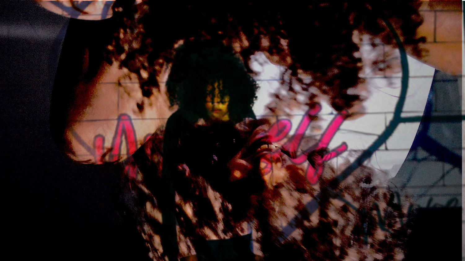 A person with curly hair stands in front of a wall with large, colorful graffiti.