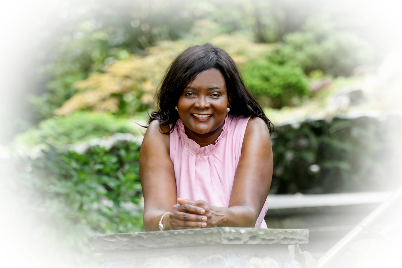 Woman smiling while leaning on a stone surface in a lush garden setting.