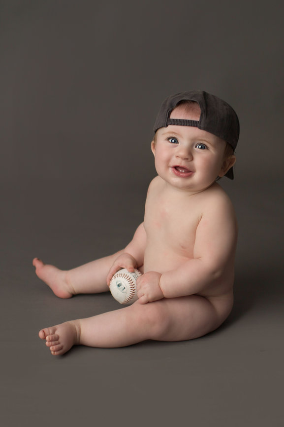 Happy baby boy with big smile sitting on grey backdrop wearing Kansas City royals hat holding a baseball during baby photo session with Kansas City, kansas baby photographer Dreamworks Photography 