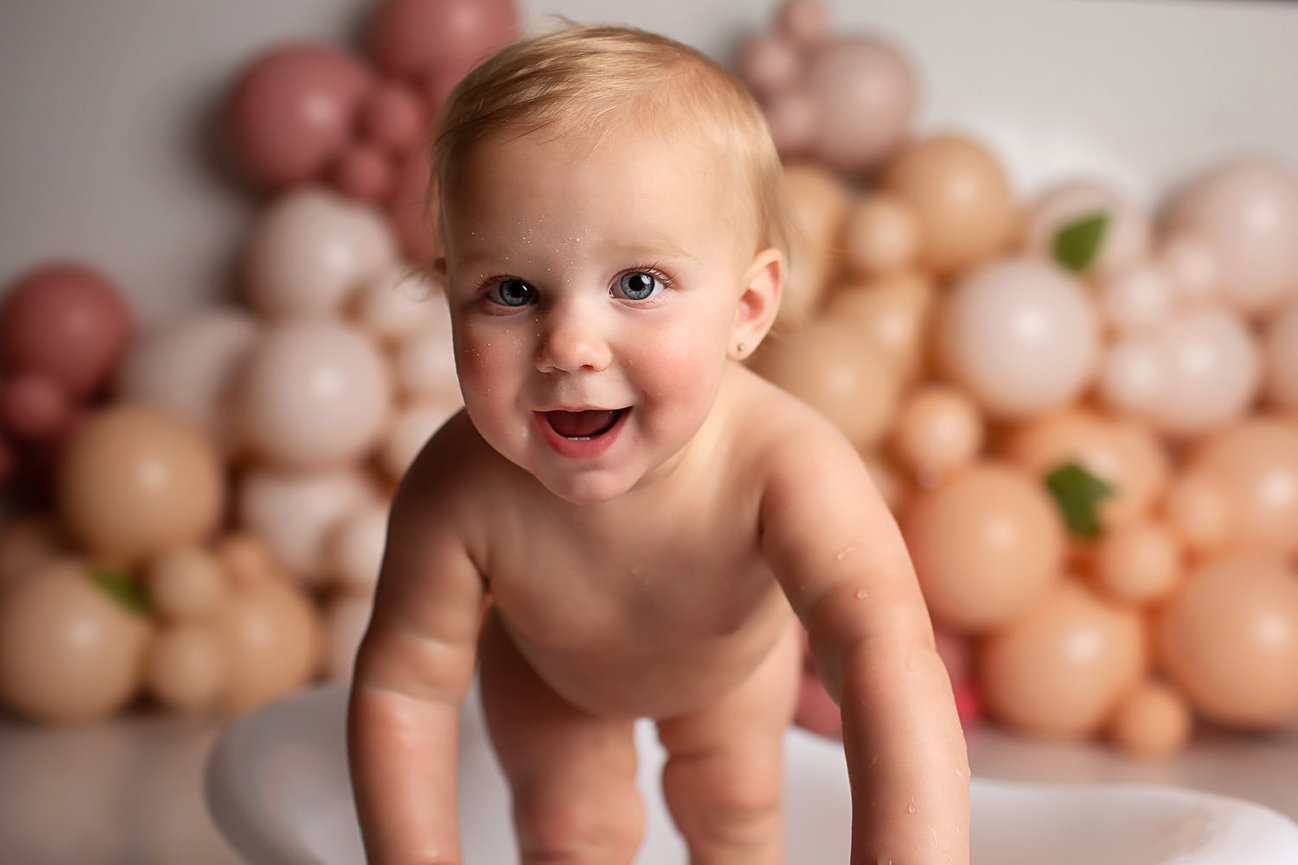 Baby girl standing and smiling in tiny bath tub during cake smash photoshoot with baby photographer Dreamworks Photography in Kansas City photography studio