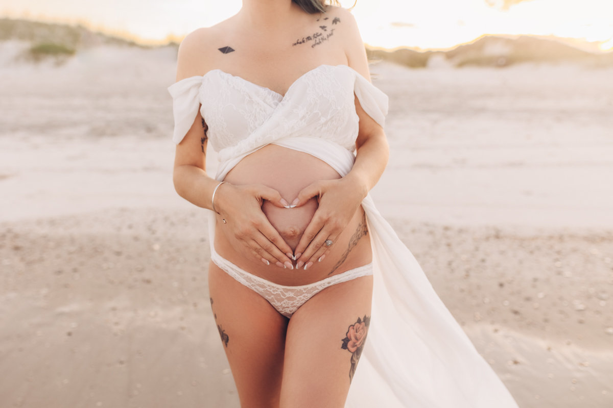 Pregnant woman in a white outfit forms a heart with hands over her belly on a beach at sunset during a maternity photography session with Catherine Whitney Photography in Jacksonville Florida