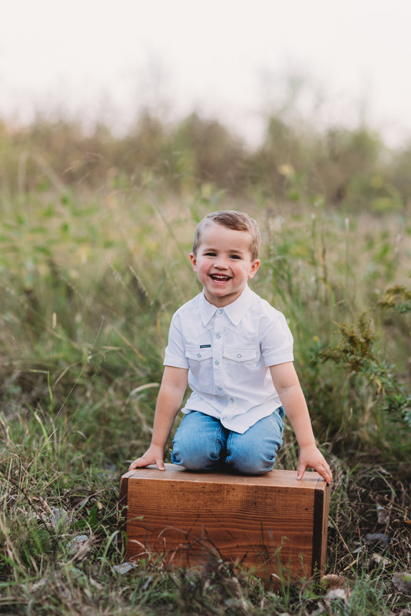 A smiling child in a white shirt and jeans sits on a wooden box in a grassy field.