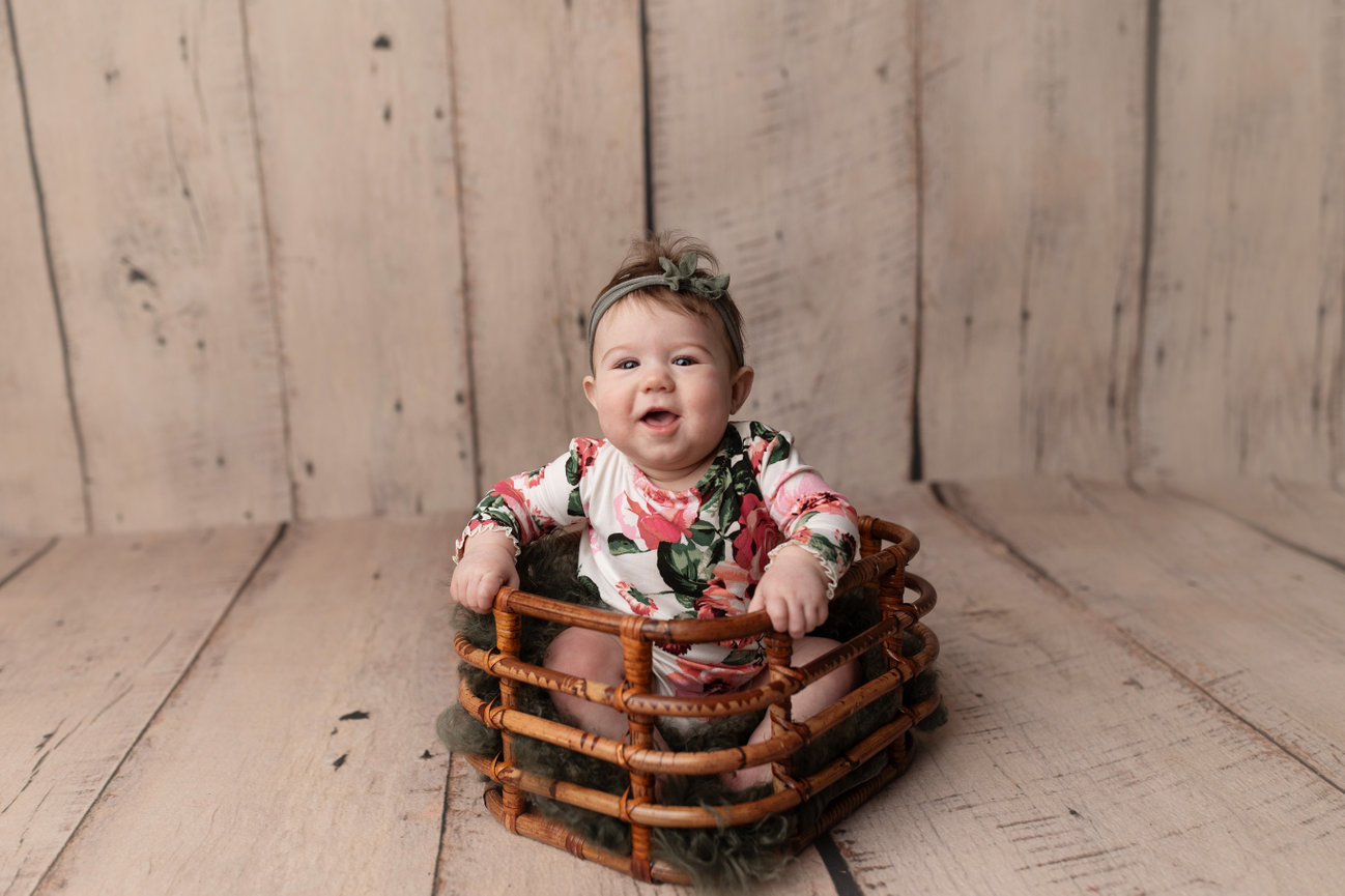 Bennington Photographer photographs baby sitting in wicker basket on wooden floor, wearing floral outfit and headband. Light wood background.