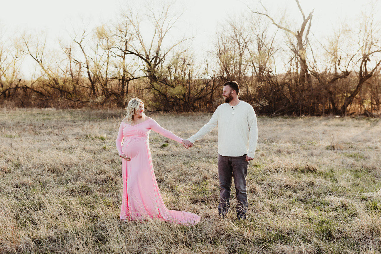 A pregnant woman in a pink dress holds hands with a man in a field at sunset.