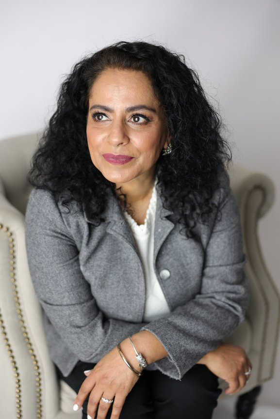 Portrait of a woman with curly black hair wearing a gray blazer, seated on a chair against a light background. authentic photos for my business and website