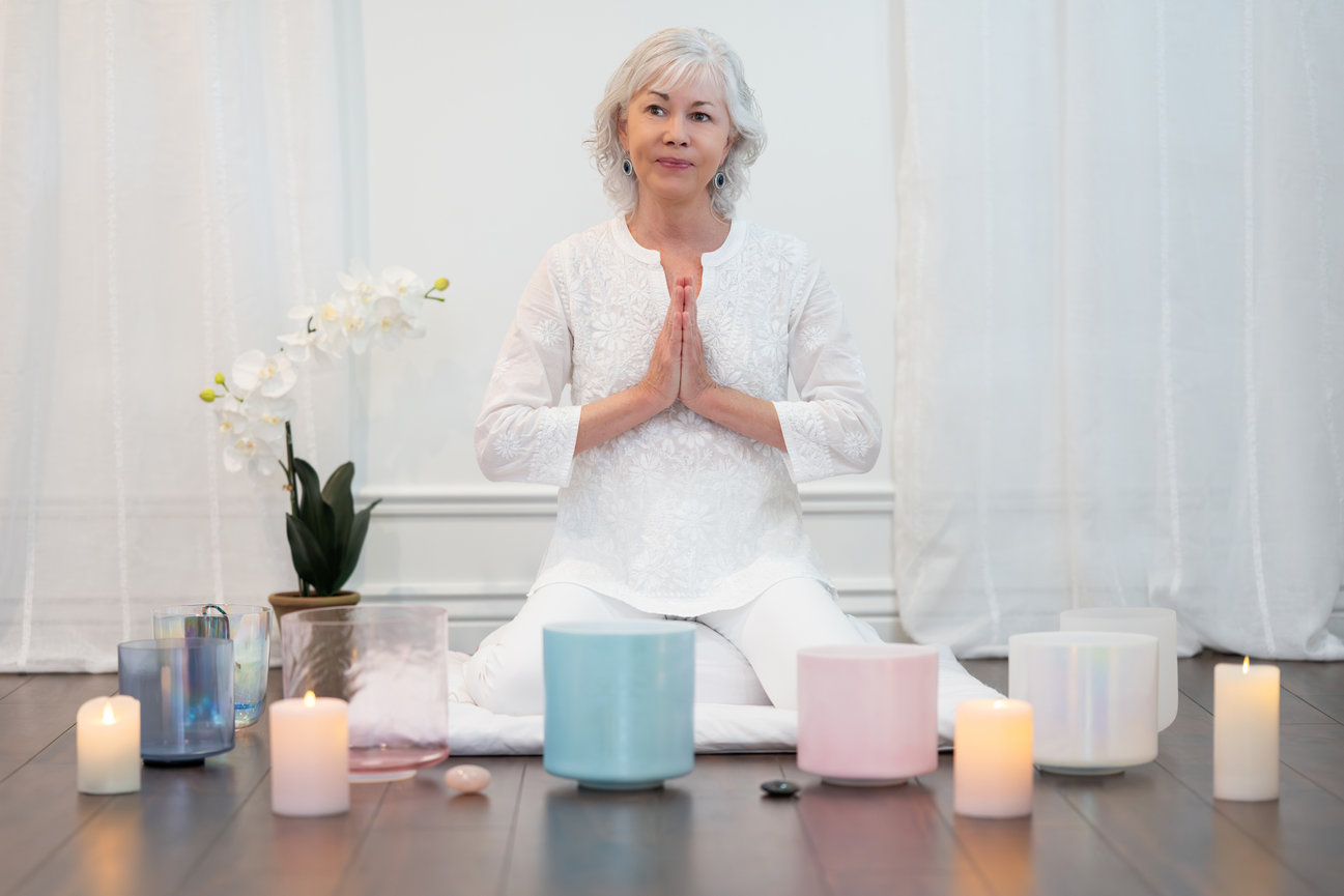 Woman sitting on ground in a prayer pose with sound bath bowls in front of her in a photo studio near Salt Lake City.
