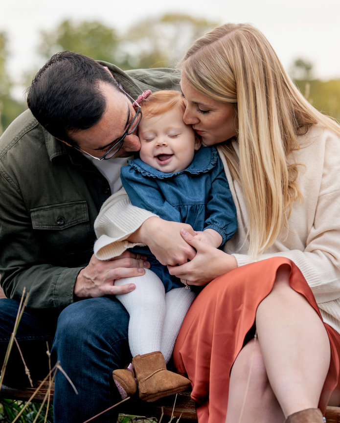 Young parents of a happy baby girl smother her with kisses while sitting on a bench in Fortville Indiana.
