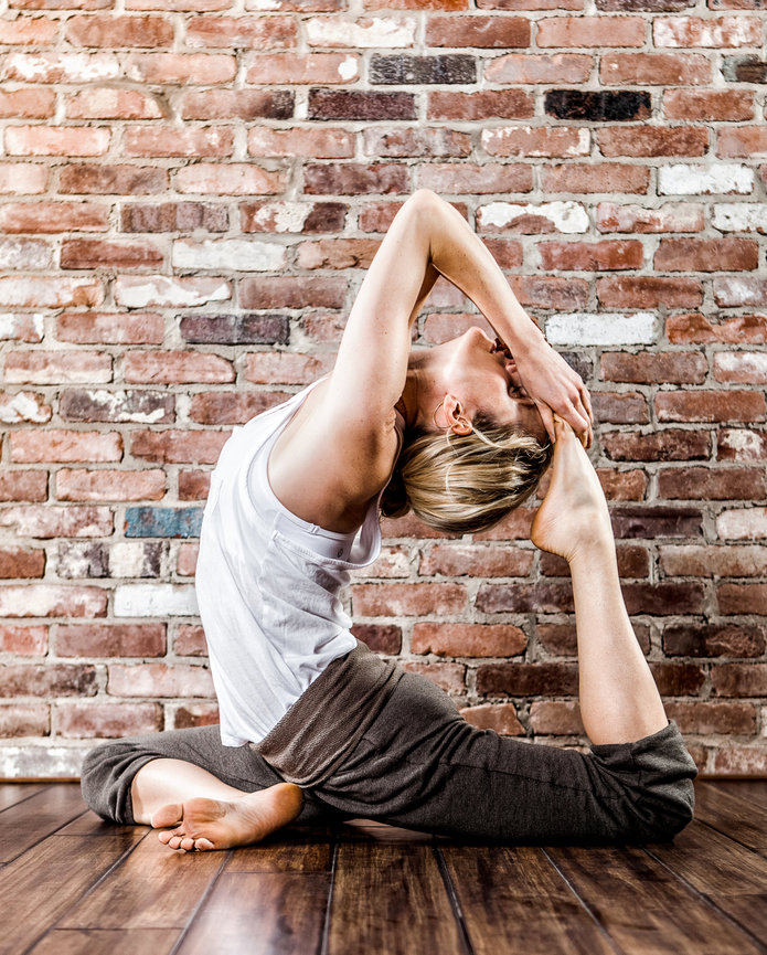 Person performing a yoga pose on a wooden floor against a brick wall, arching back with one leg bent and hands holding the foot.