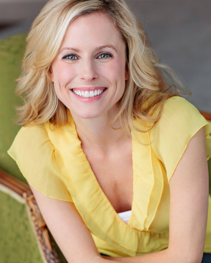 Smiling woman with blonde hair wearing a yellow blouse and bracelets, sitting on a green chair, looking up.