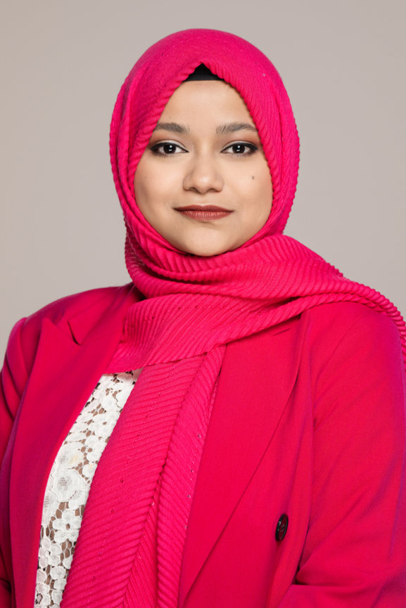 A woman wearing a pink hijab and coat, posing with a neutral expression against a light background.