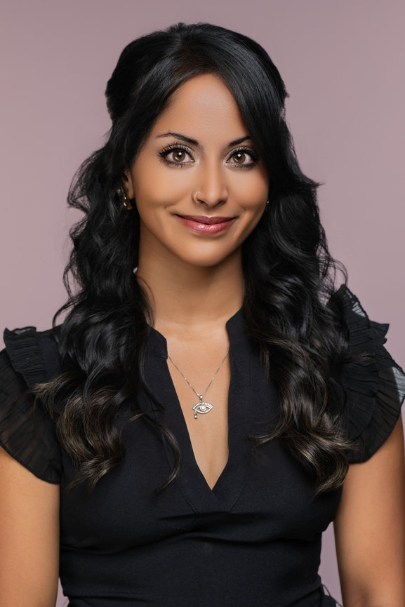 Portrait of a woman with long, wavy hair, wearing a black top and a silver necklace, against a pink backdrop.