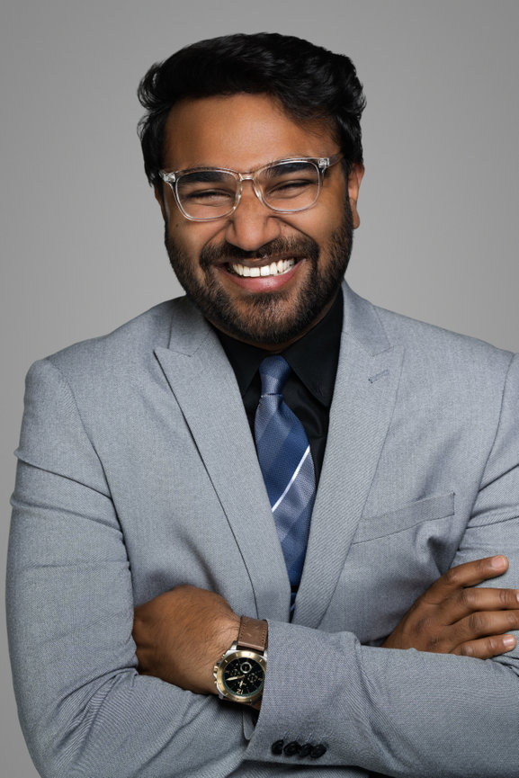 Smiling man in a gray suit with glasses, arms crossed, against a neutral background.