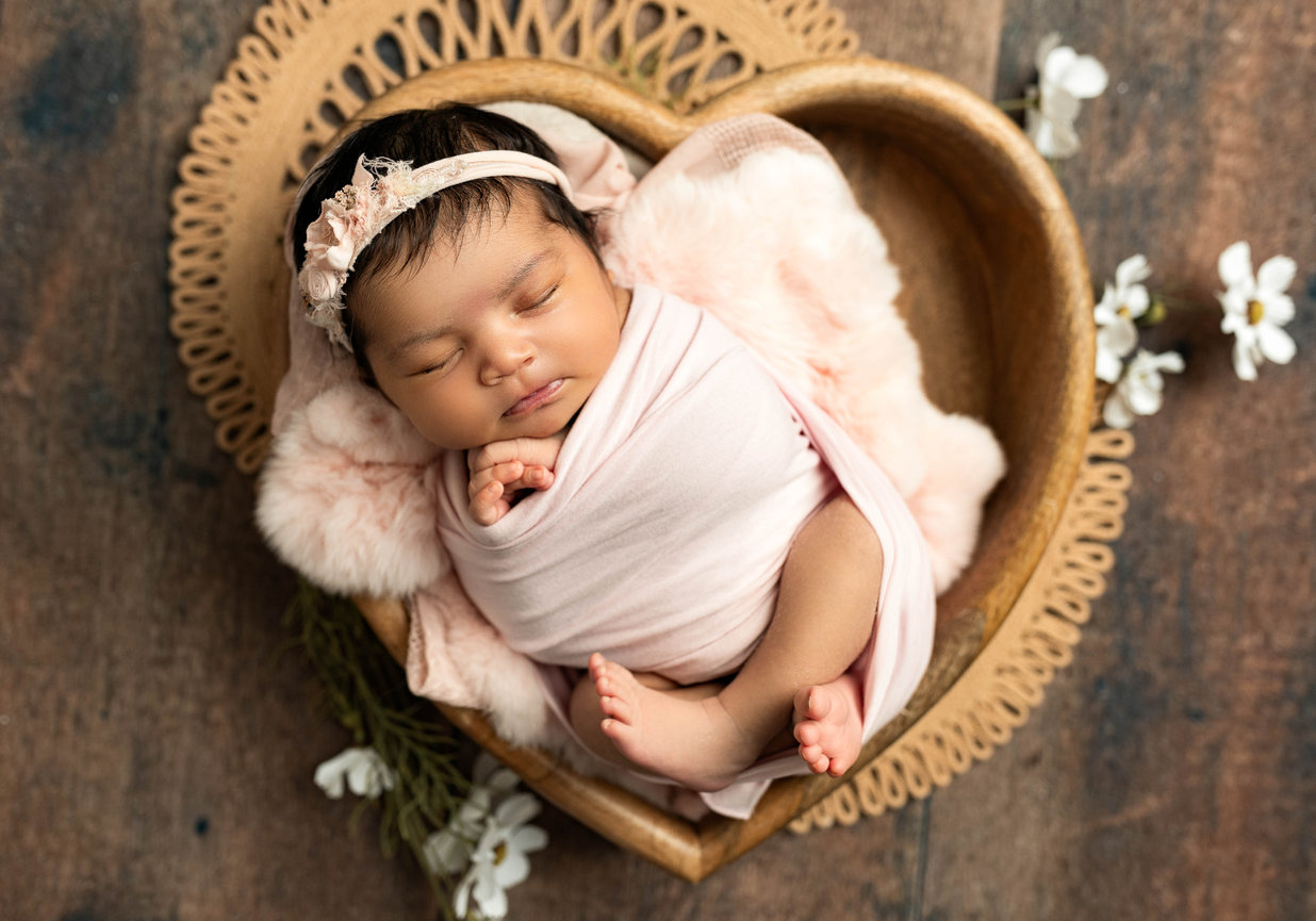 Newborn girl with pink headband swaddled in heart basket