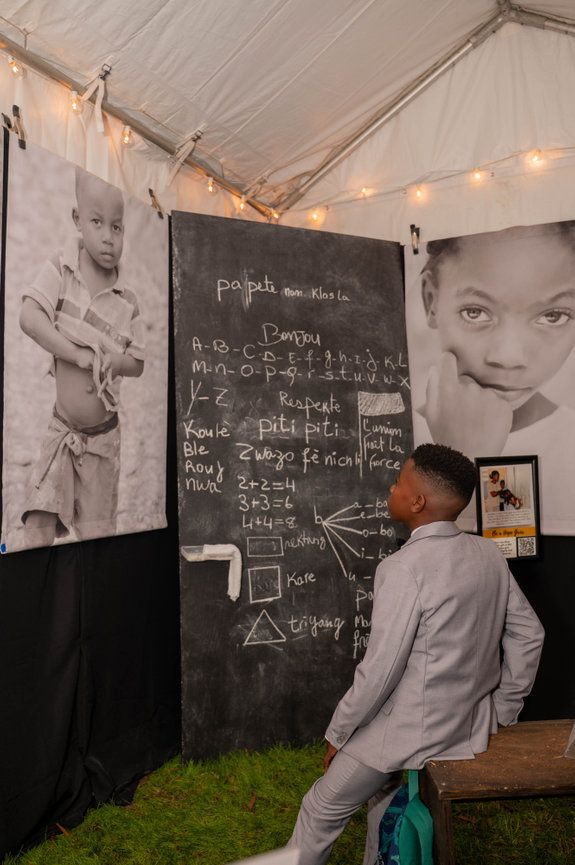 Child in suit viewing a chalkboard with math equations and Creole words under black-and-white portraits in a makeshift Haitian classroom captured by Jo Hayes Images Charlotte Event Photography