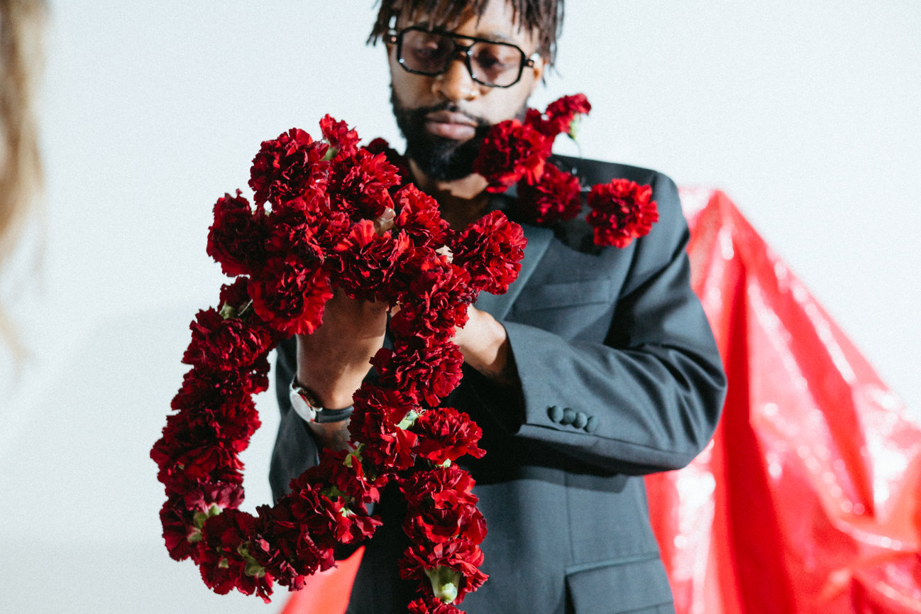 Man in a suit holding a red floral garland, with a red draped background.