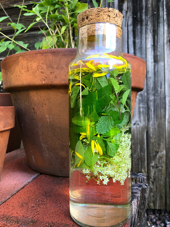 Corked glass bottle filled with herbs and flowers, placed in front of clay pots.