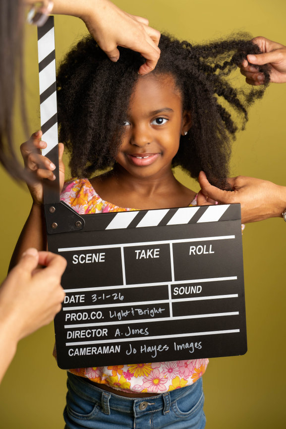 Young girl smiling, holding a clapperboard in front of an olive green background, with hands styling her hair captured by cameraman Jo Hayes Images