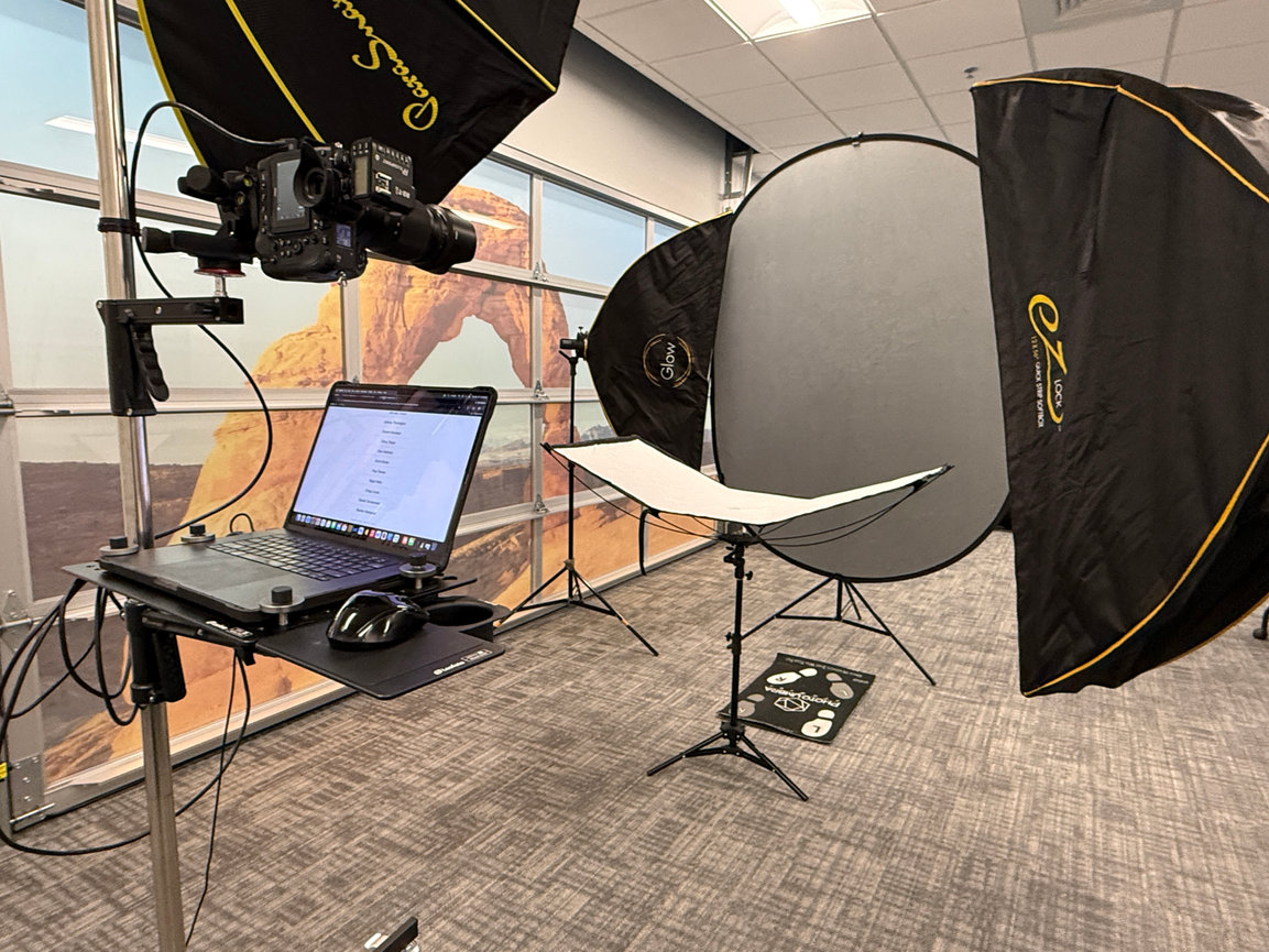 Photography studio setup with camera, lights, laptop on stand, and backdrop featuring a desert arch scene.