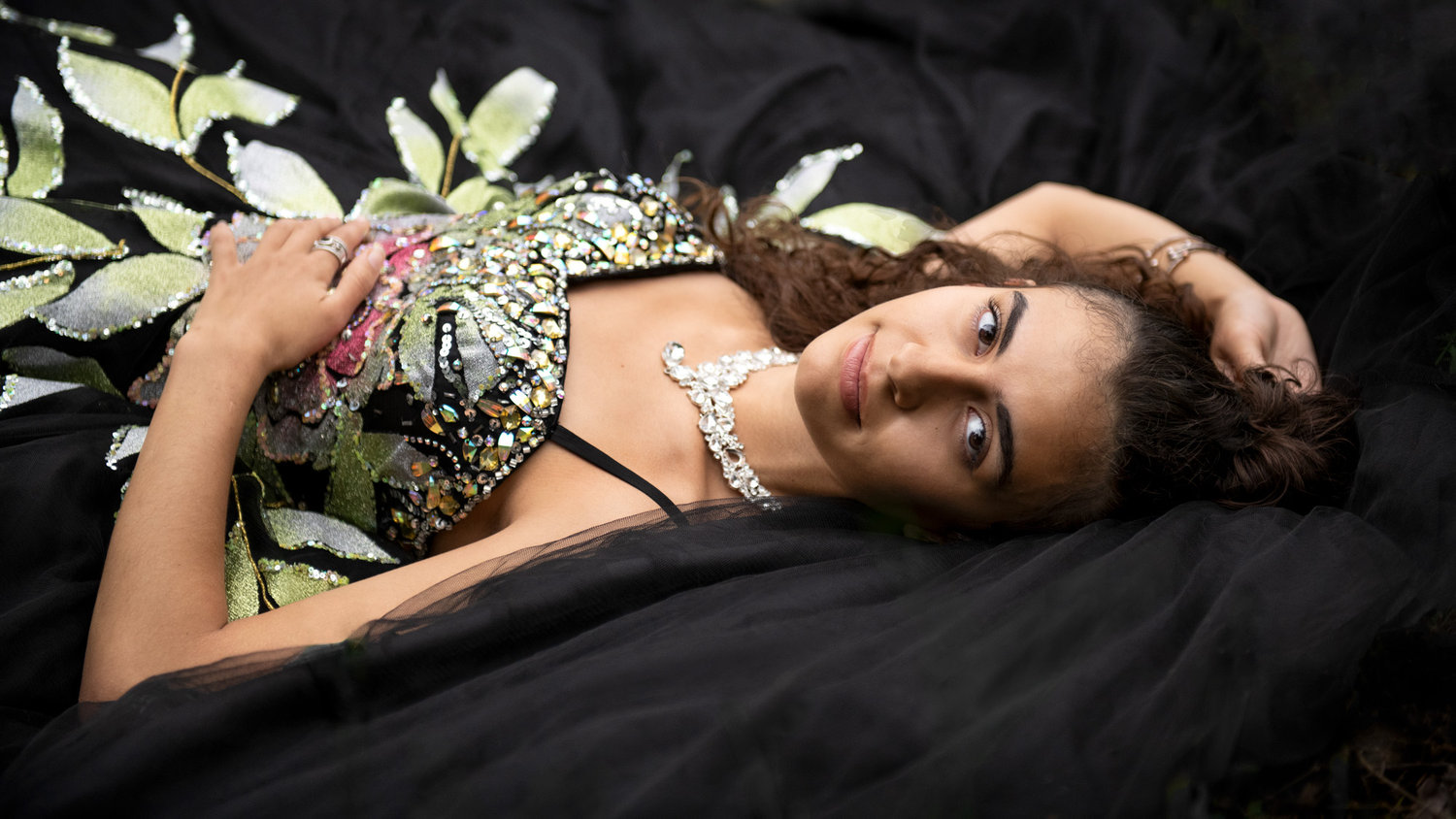 Portrait of a female high school graduate in her jeweled gown reclining on black fabric, wearing a sparkling necklace and looking at the camera.