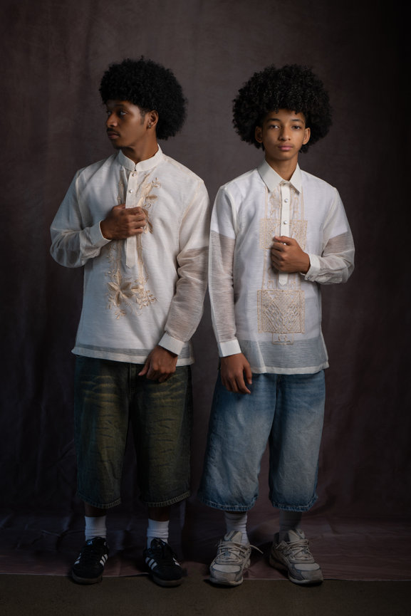 Two young men in embroidered white shirts and denim shorts pose against a dark studio backdrop.