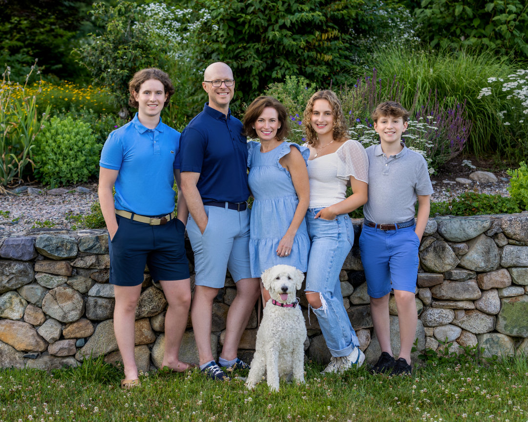 outdoor family portrait in blue of three teenaged children and mother and father and family dog by stone wall and garden canton ma