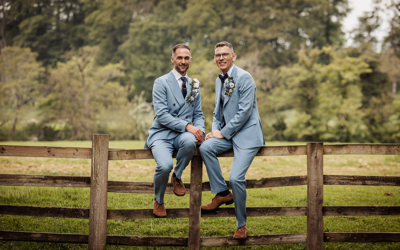 Two men in light blue suits sit on a wooden fence outdoors in a green field