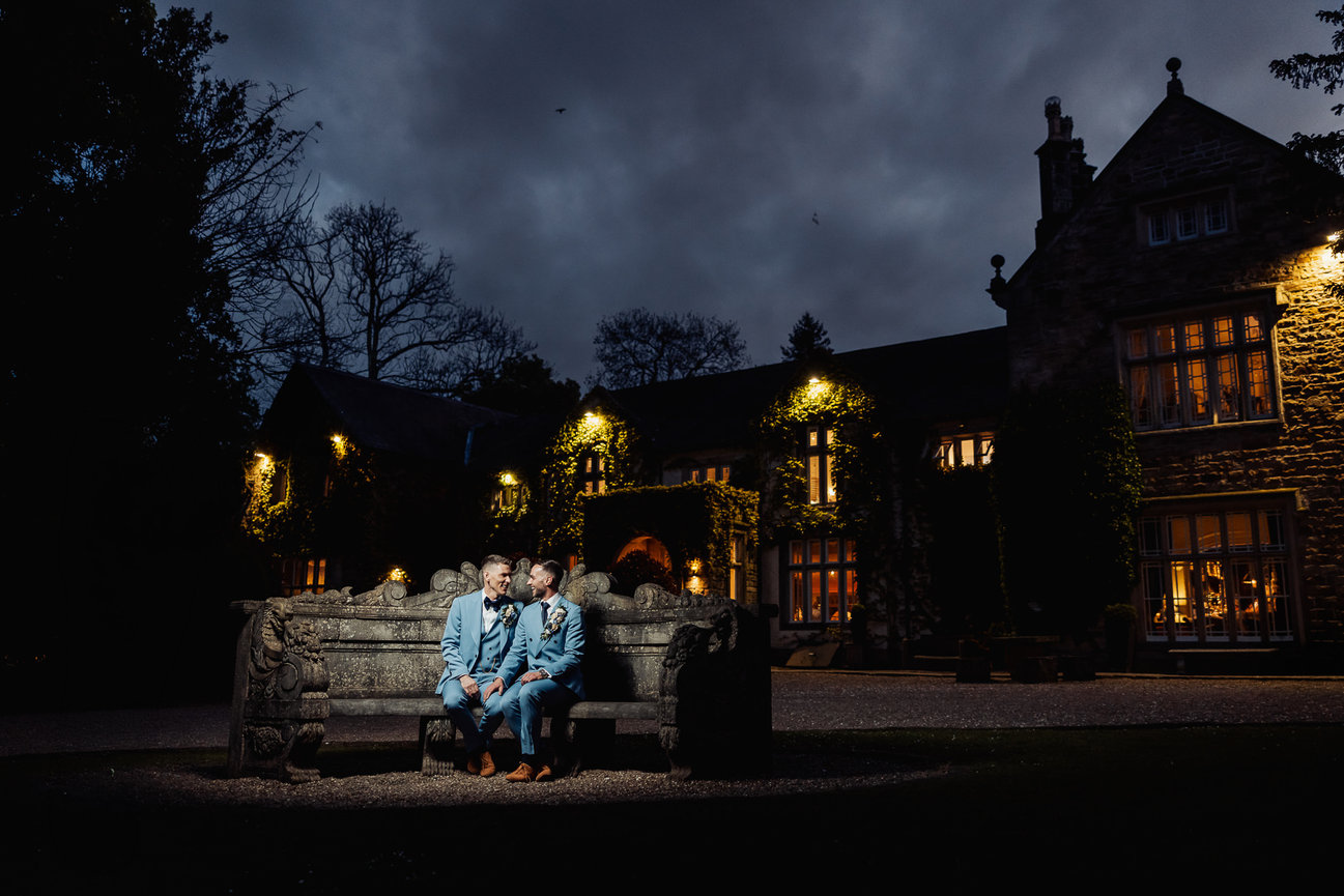 Two men in blue suits sit on a stone bench outside a lit manor at night