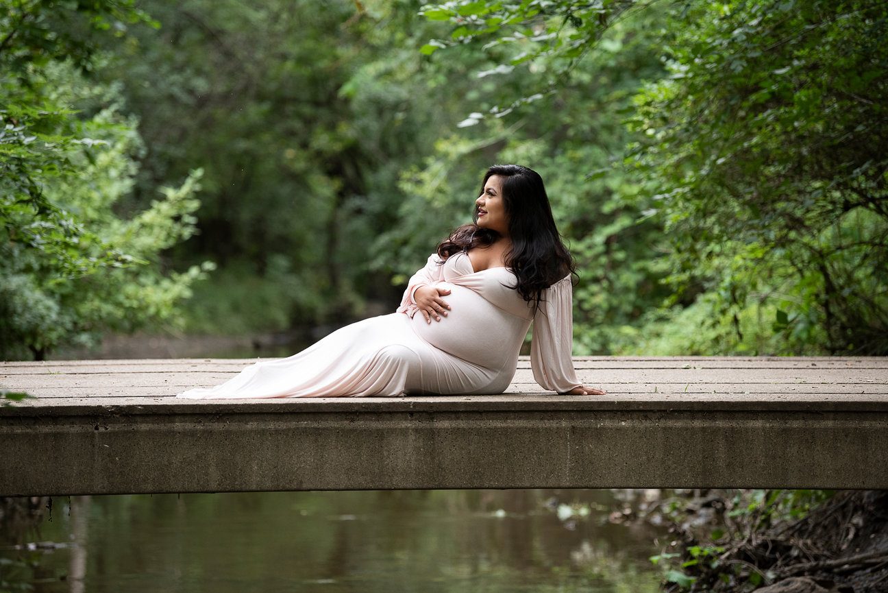 A pregnant woman in a white dress sits on a bridge, surrounded by lush green trees and a stream below. She is gazing to the side with a serene expression, gently holding her belly.