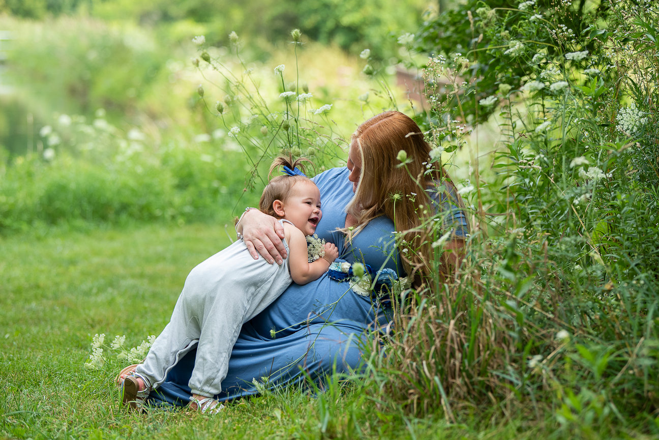 A woman with long red hair, wearing a blue dress, sits on grass near a bush. She hugs a laughing child in a white outfit. The scene is surrounded by lush greenery, and the woman has a joyful, content expression.