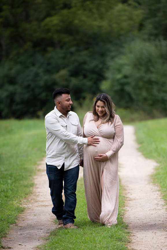 A couple strolls hand in hand along a path embraced by lush greenery. The man gently touches the womans pregnant belly, celebrating this beautiful journey of pregnancy with smiles, both dressed in casual outfits.