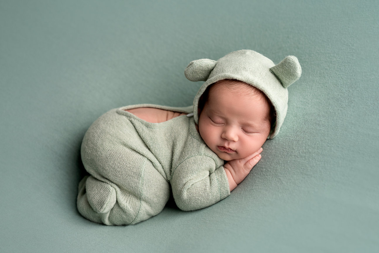 A sleeping baby dressed in a soft, light green outfit with bear ears on the hood lies peacefully on a matching surface. Curled up with hands under the chin, this serene scene could easily be set in charming Lake Zurich, Illinois.