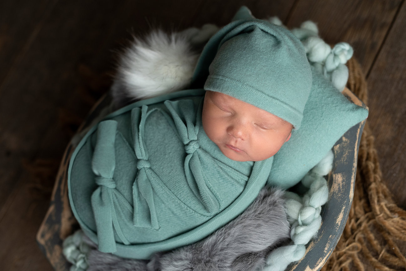 In a cozy setting in Roselle, a sleeping newborn baby is swaddled in a soft teal wrap with a matching hat. The little one rests on a bed adorned with furry grey accents and braided fabric, set against a rustic wooden backdrop.