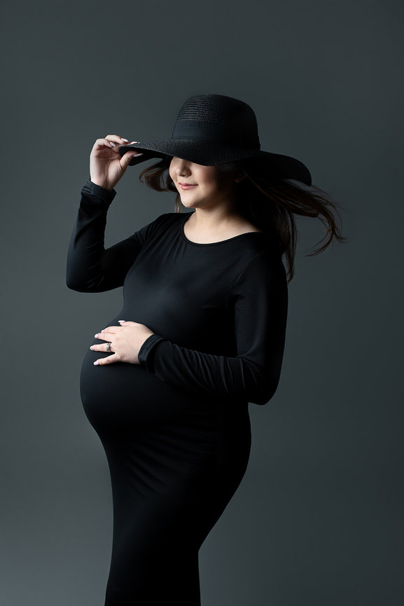 A pregnant woman in a fitted black dress and wide-brimmed hat poses gracefully against a gray background. As she touches her hat and cradles her belly, thinking of the baby care classes near me, her hair flows slightly, capturing a serene moment.