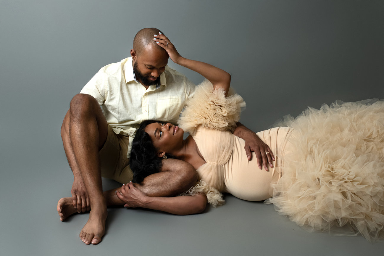 In a serene studio setting, a couple basks in tender moments. The woman, in a flowing beige tulle dress, rests her head on her husbands lap as he gently places his hand on her head. A maternity photographer captures the love.