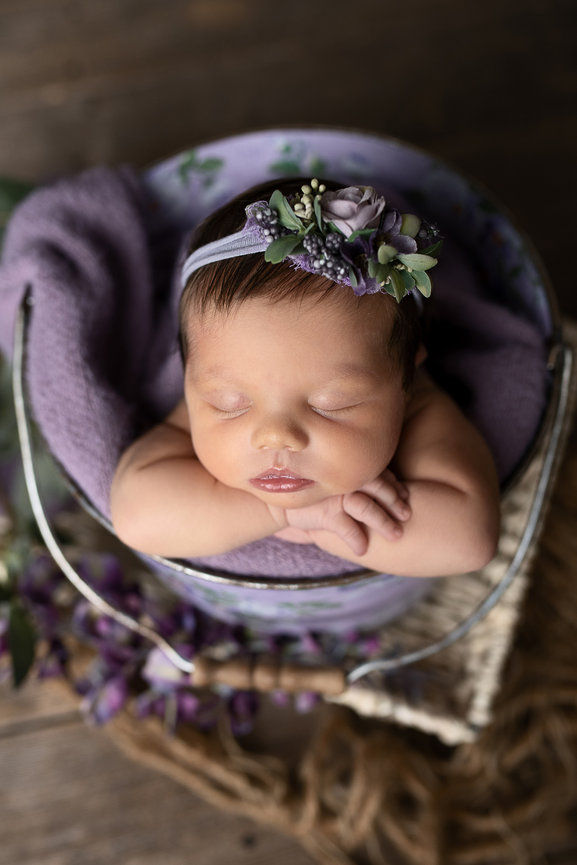 Newborn baby sleeps peacefully in a floral-decorated bucket, embodying the serene essence of Crystal Lake. The baby, wearing a headband with purple flowers, rests on a purple blanket. The scene unfolds on a wooden surface with purple flowers nearby.