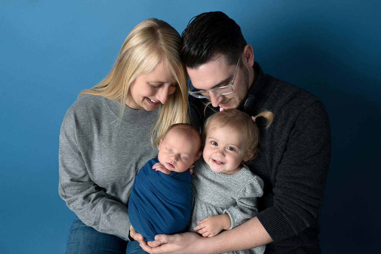 A family sits together against a blue background, embodying the warmth of Winfield, Illinois. The mother and father are on either side, holding a newborn swaddled in blue and a smiling toddler. Cozy sweaters enhance the warm and loving atmosphere.