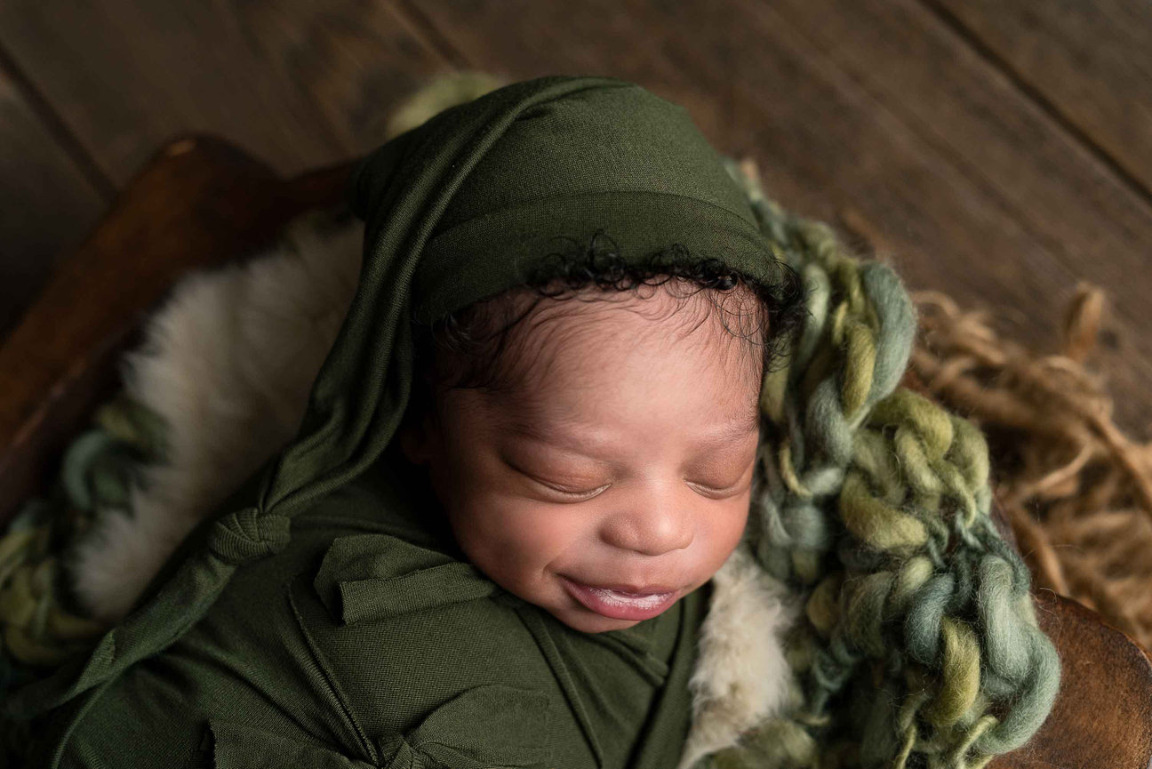 A newborn baby wrapped in an olive green swaddle and hood sleeps peacefully on a textured blanket. The cozy setting, reminiscent of a serene corner in Almora Heights, Illinois, is enhanced by the warm tones of the wooden floor.