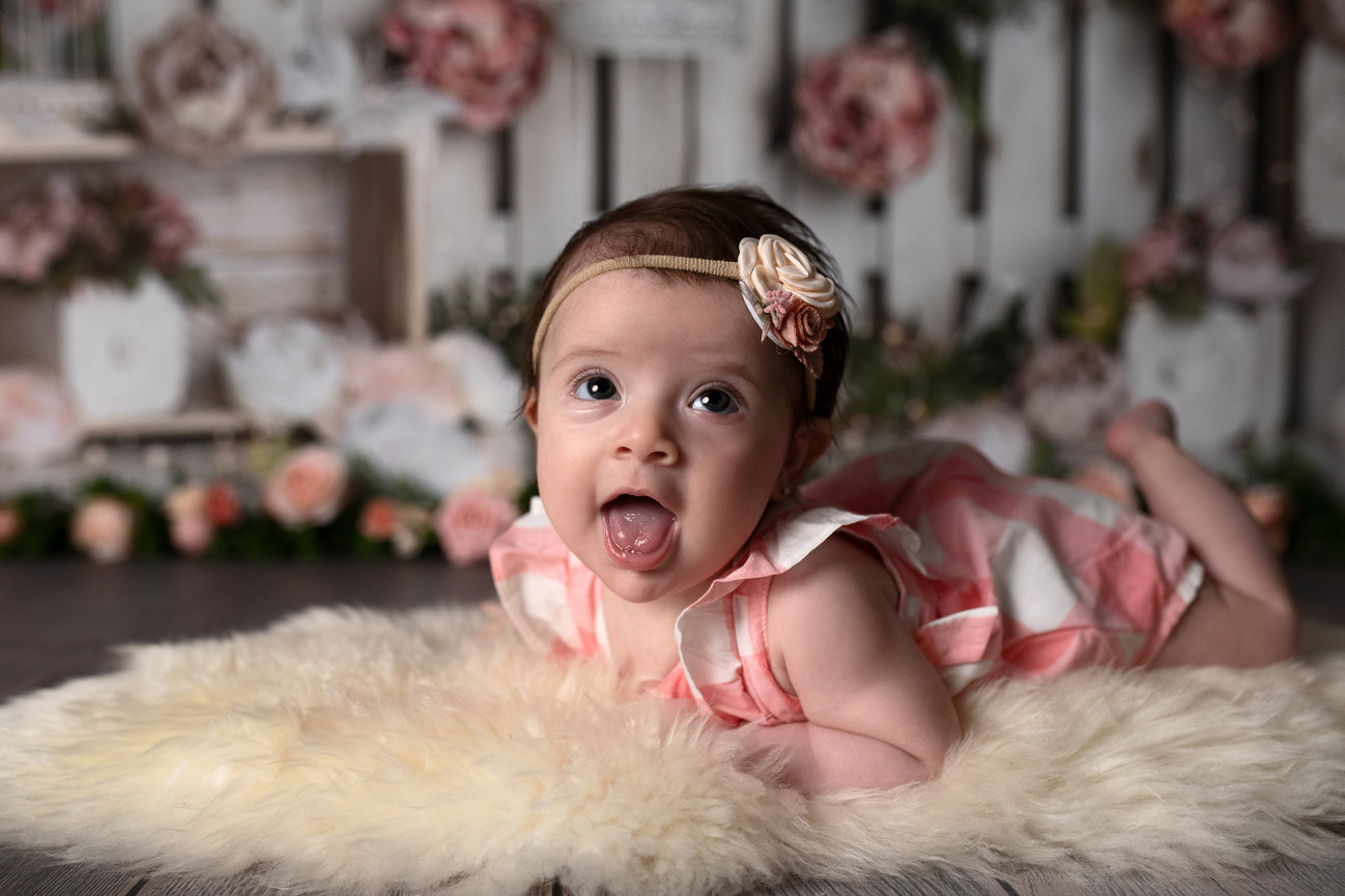 A baby wearing a pink dress and a floral headband lies on a fluffy rug, surrounded by soft pink and white flowers. Captured skillfully by a photographer near me, the baby gazes at the camera with their tongue playfully out.