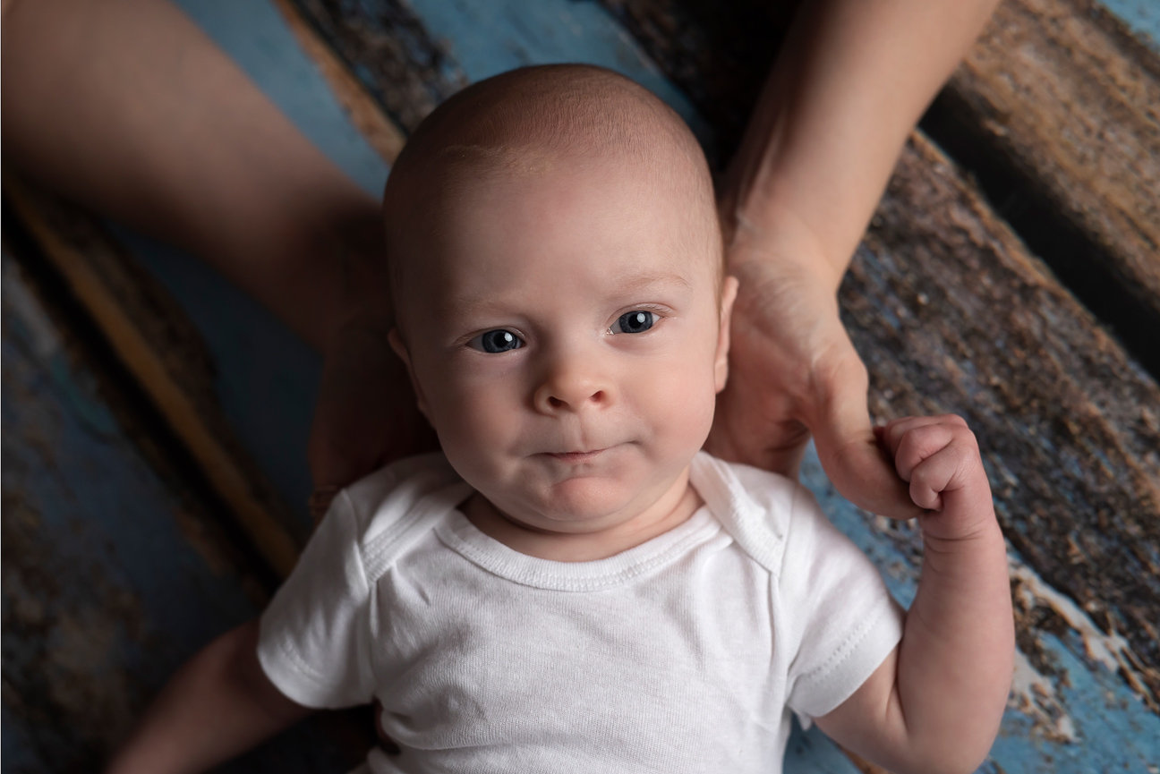 A baby with blue eyes and a slight smile lies on a wooden surface, wearing a white onesie. An adults hands gently support the babys head from behind. Capturing the essence of baby photography near me, a textured blue and brown pattern sets.