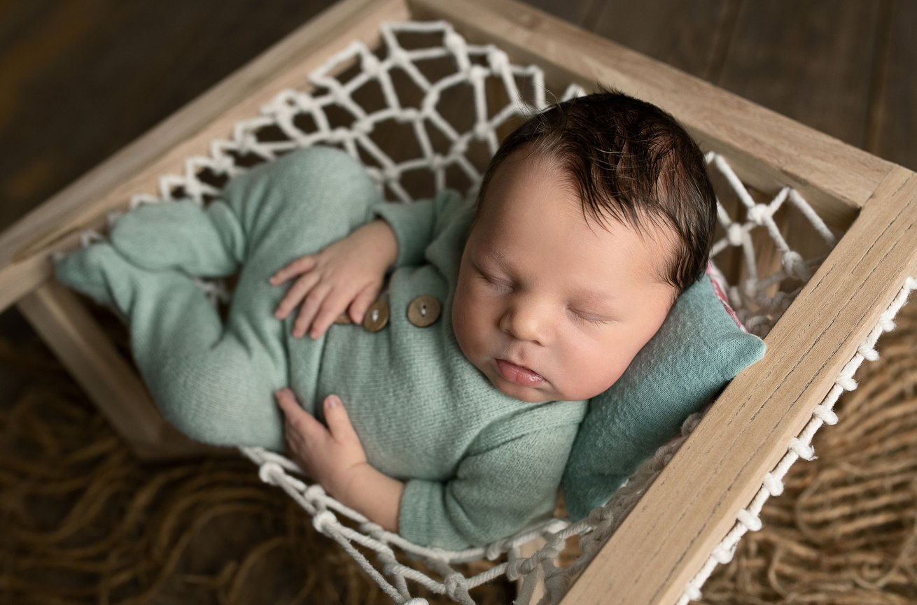 A sleeping baby in a green onesie rests in a wooden-framed net hammock, with a matching green pillow under their head. The serene scene unfolds on a wooden floor in St. Charles, Illinois, complemented by a soft, brown textured blanket underneath.