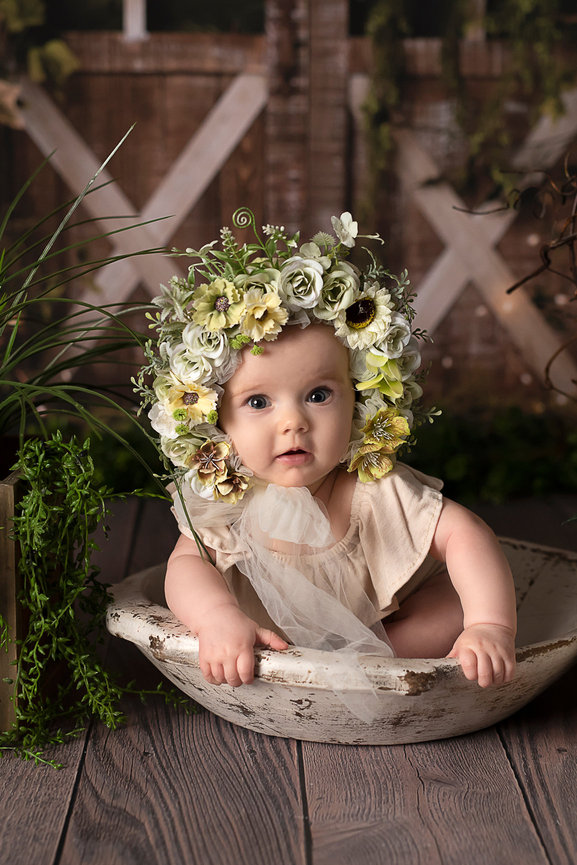 A talented photographer captures a baby wearing a floral crown, sitting in a rustic wooden bowl. The child is dressed in light beige, surrounded by greenery. In the background, a wooden fence with vines completes this natural and serene setting.