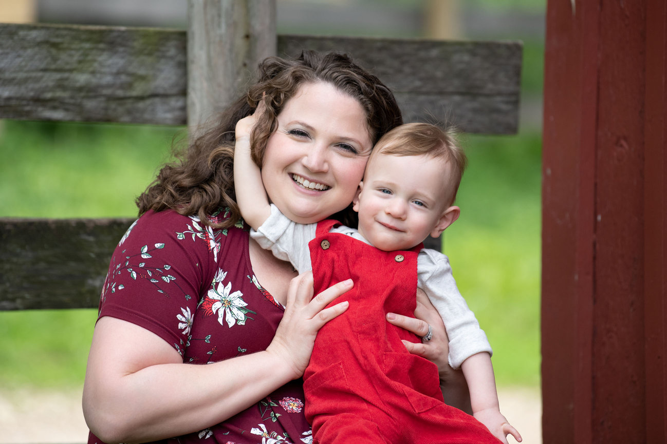 A woman with curly hair and a floral maroon top smiles while holding a small child in red overalls. Captured by a talented family photographer in Elgin, they sit outdoors in front of a wooden fence, surrounded by lush greenery.