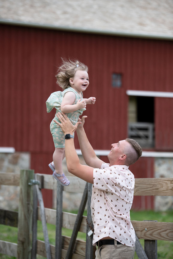 In a delightful capture of Schaumburg, IL family photography, a man joyfully lifts a smiling toddler above his head. The child wears a light green outfit adorned with butterflies, while the man sports a polka dot shirt and glasses.